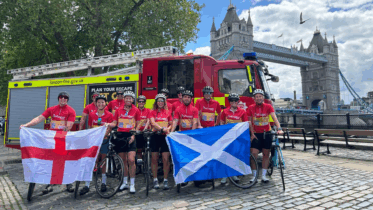 A group of cyclists in red jerseys pose with England and Scotland flags in front of a fire truck, with Tower Bridge and London’s skyline visible in the background.