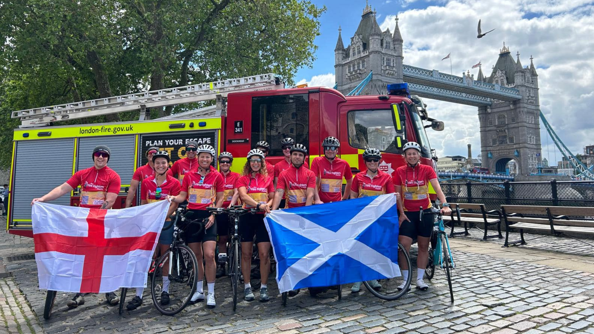 A group of cyclists in red jerseys pose with England and Scotland flags in front of a fire truck, with Tower Bridge and London’s skyline visible in the background.