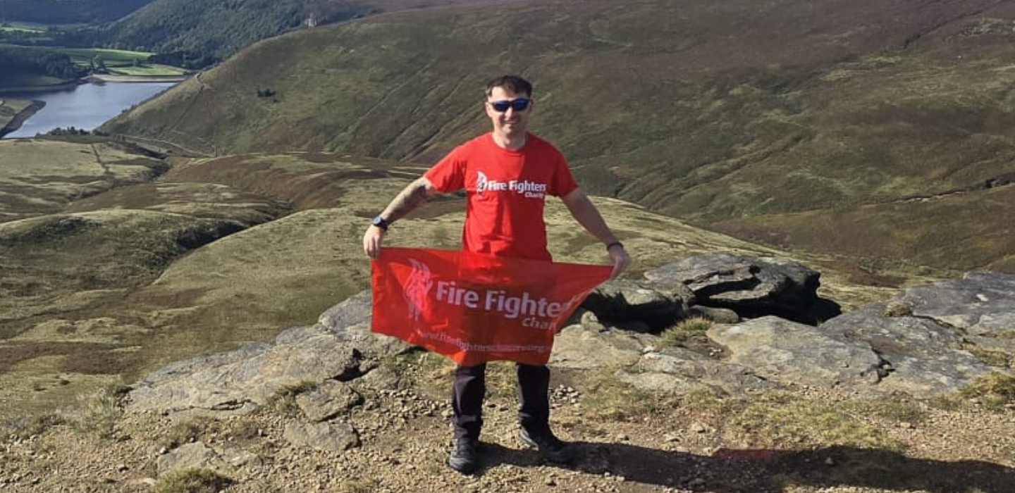A man in a red “Fire Fighter” shirt holds a matching fire fighter charity banner while standing on a rocky hilltop with green valleys and a winding river in the background.