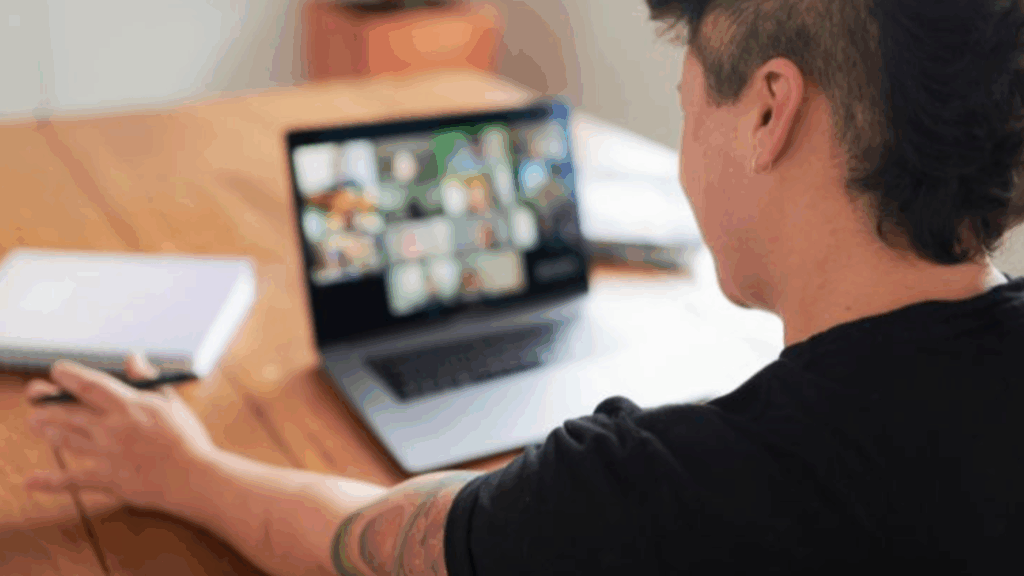 A person with a tattooed arm sits at a wooden table, looking at a laptop showing a grid of people in a video call. A notebook and pen are nearby.