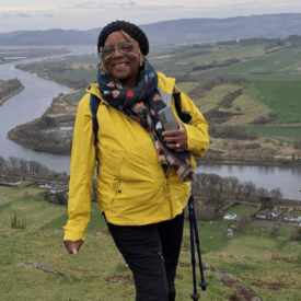 A woman wearing a yellow jacket and glasses smiles while hiking on a hillside, holding walking poles. A winding river and green fields stretch out in the valley behind her under a cloudy sky.