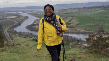 A woman wearing a yellow jacket and glasses smiles while hiking on a hillside, holding walking poles. A winding river and green fields stretch out in the valley behind her under a cloudy sky.