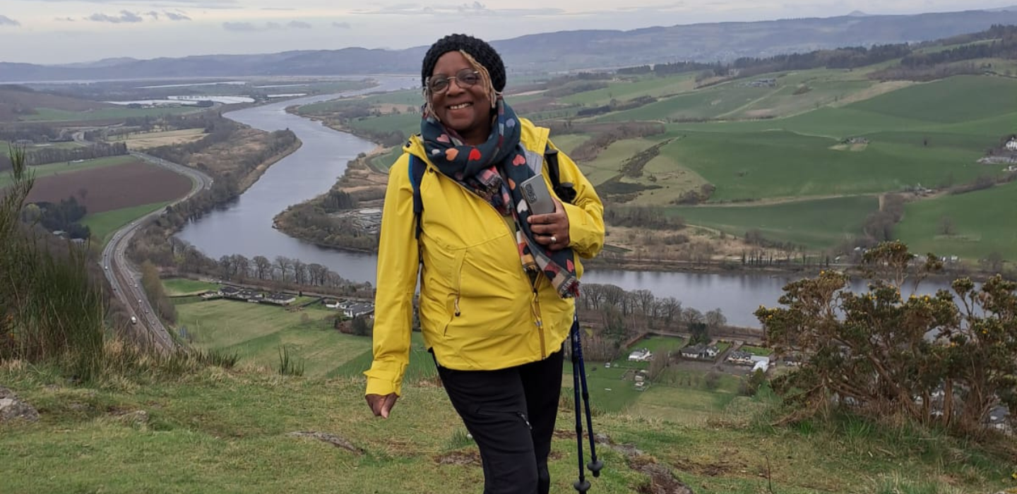 A woman wearing a yellow jacket and glasses smiles while hiking on a hillside, holding walking poles. A winding river and green fields stretch out in the valley behind her under a cloudy sky.