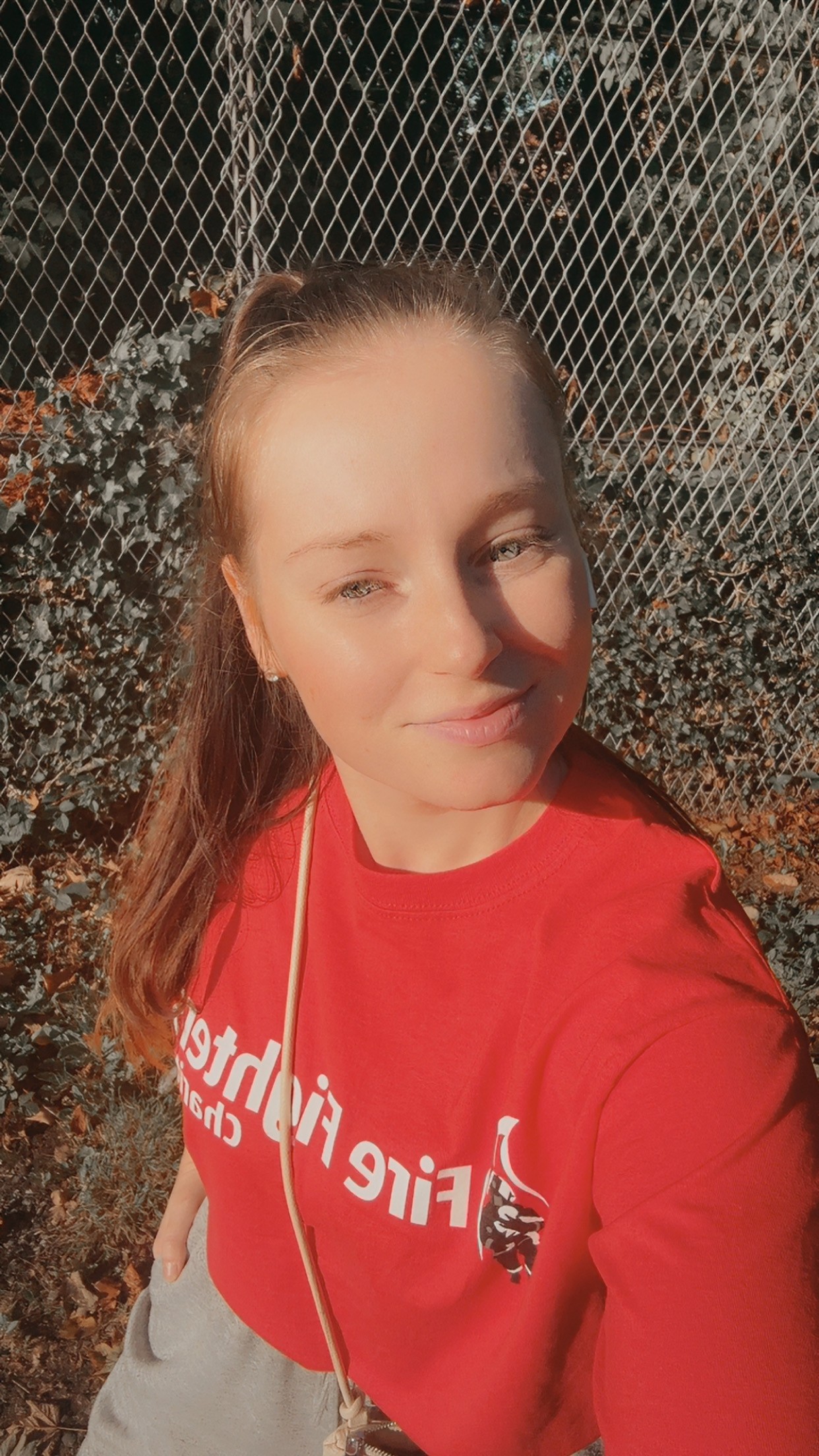 A woman with long brown hair in a ponytail takes a selfie outdoors, smiling and wearing a red "Fire Fighter" t-shirt. There's a chain-link fence and foliage in the background.