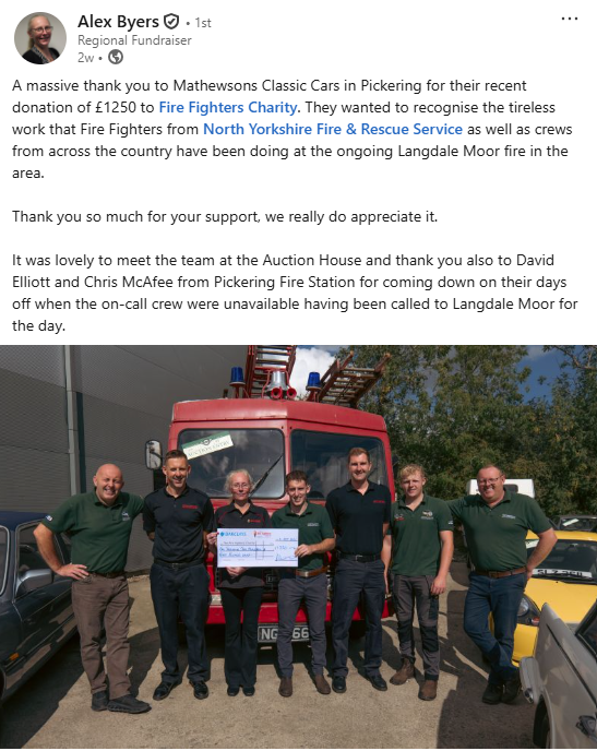 A group of seven people stand smiling in front of a red fire truck, holding a large cheque for £1,250. Two men wear uniforms. The setting appears to be outside a fire station on a sunny day.