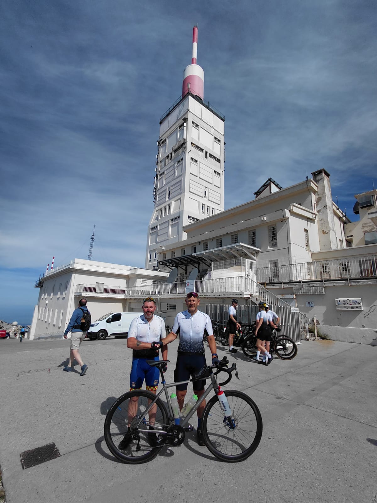 Two cyclists in matching jerseys stand with their bikes in front of the iconic white tower of Mont Ventoux under a blue sky, with other people and cyclists nearby.