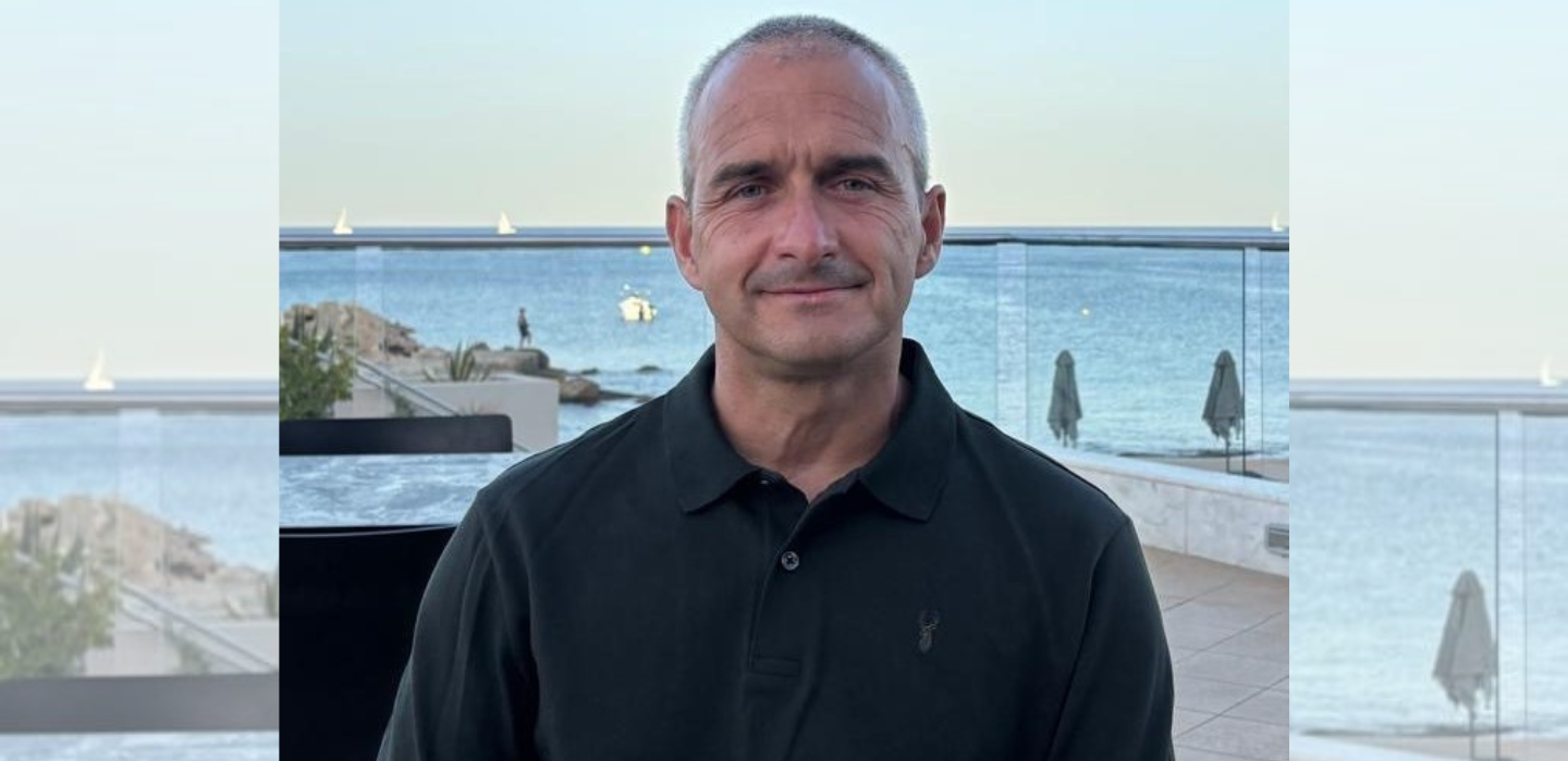 A man with short gray hair wearing a black polo shirt sits outdoors in front of a glass railing, with an ocean and beach scene in the background. The sky is clear and there are a few umbrellas and boats visible.