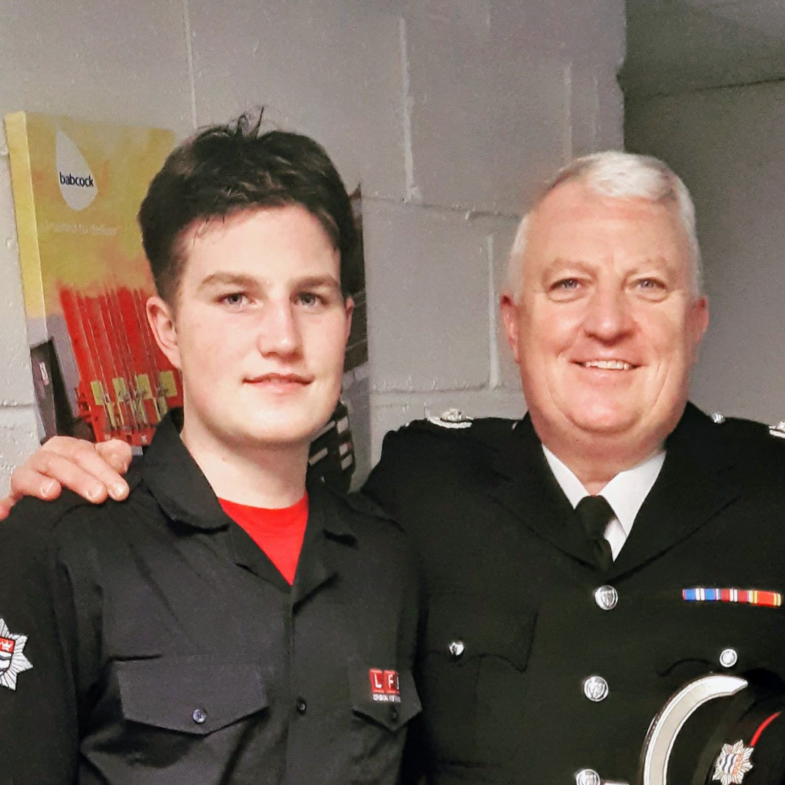 Two men in uniform stand side by side, smiling at the camera. One is younger and wearing a dark shirt with badges, while the older man has gray hair and is dressed in a black formal jacket with medals and insignia.