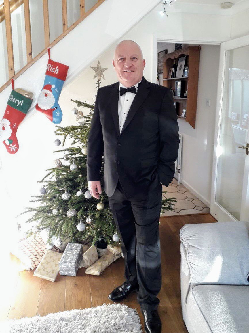 A man in a black tuxedo stands smiling by a decorated Christmas tree with presents underneath. Two stockings labeled "Cheryl" and "Rich" hang on a staircase in a bright, cozy living room.