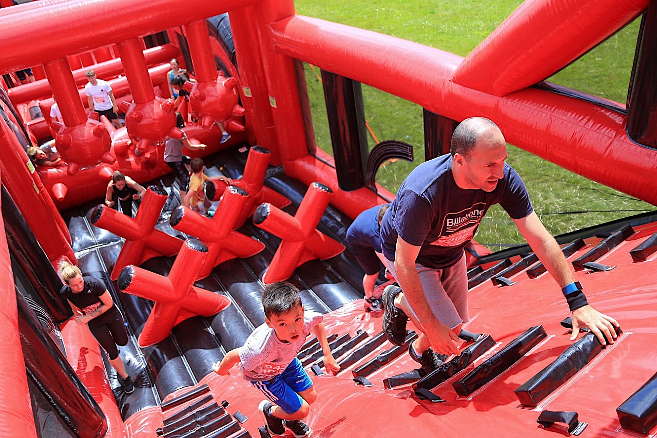 Adults and children climb a large red and black inflatable obstacle course outdoors on a grassy field, with others navigating obstacles in the background.