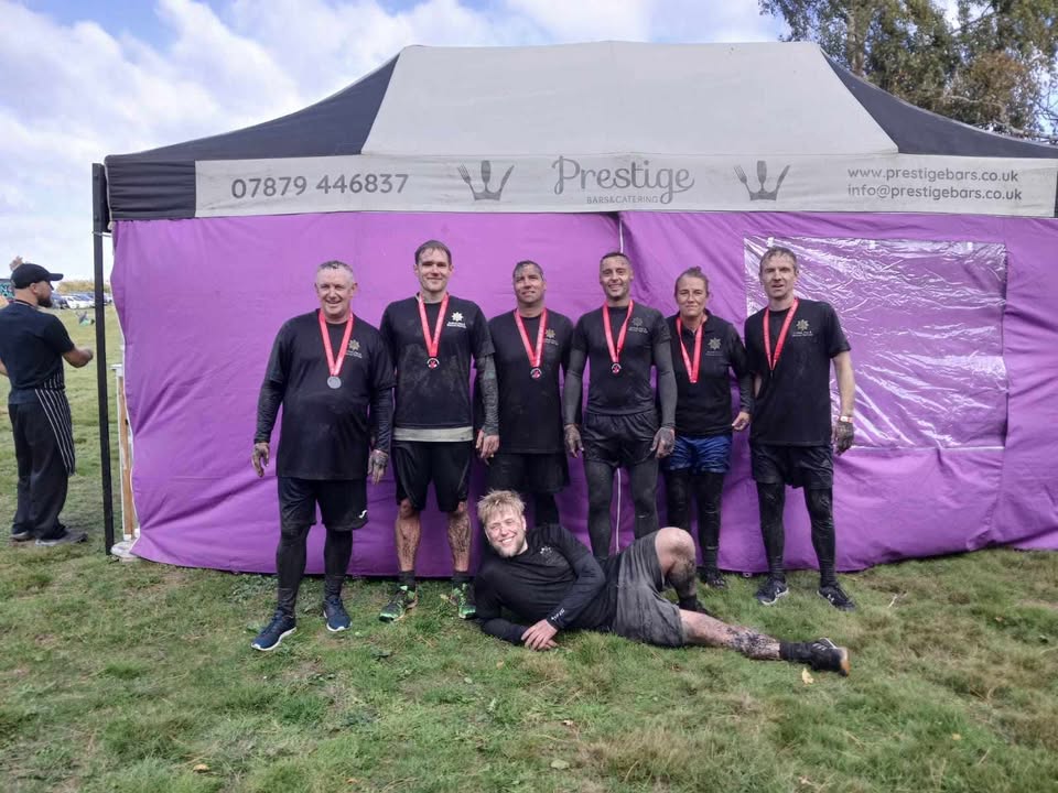 Seven men wearing black athletic clothes and red medals pose in front of a purple tent, with one man lying on the grass in front, all showing signs of mud from an outdoor event.