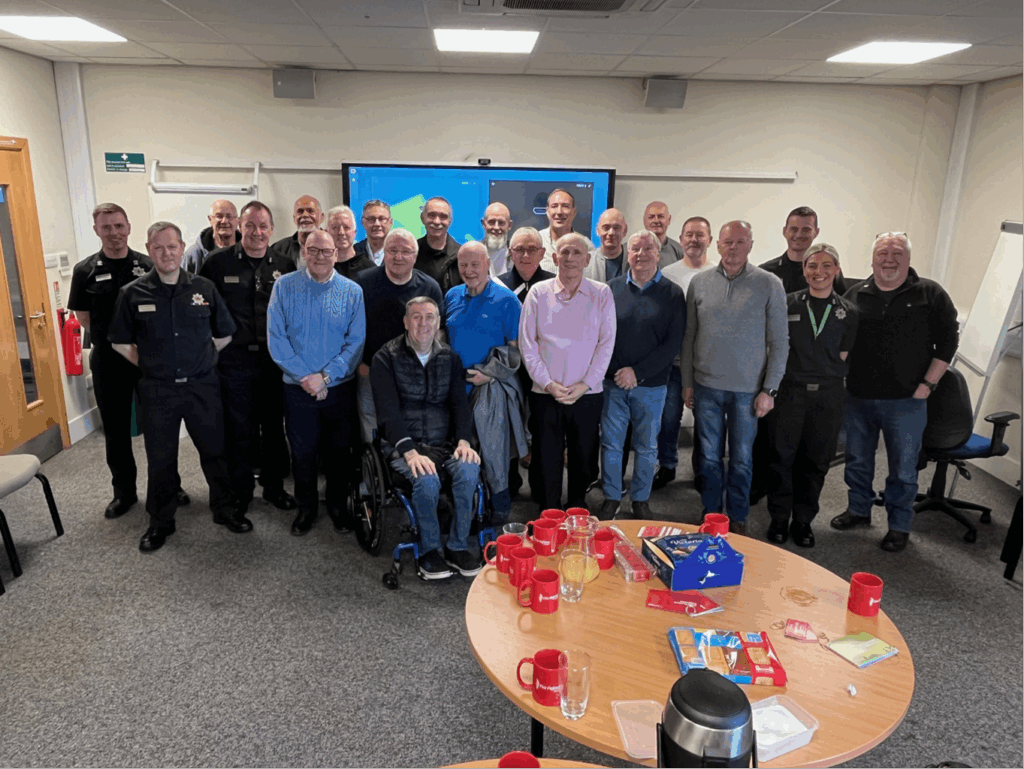 A group of about 25 people, mostly men, pose and smile together in a meeting room. A round table with red cups, snacks, and a coffee pot is in the foreground. A screen and flip chart are visible in the background.