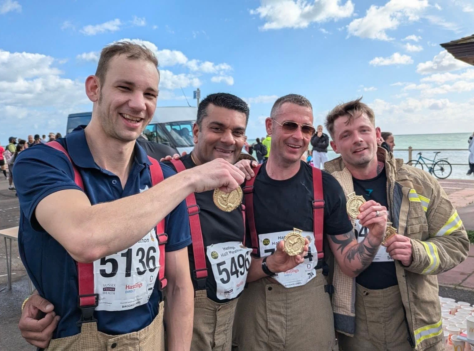 Four smiling men in firefighter gear and race bibs proudly hold medals at an outdoor event by the sea, with a cloudy sky and other people in the background.