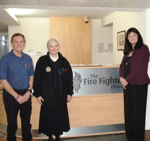 Four people stand and smile in front of a reception desk with a sign that reads "The Fire Fighters Charity." The background includes a wooden counter and light-colored walls.