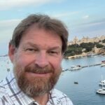 A bearded man in a plaid shirt smiles at the camera with a scenic waterfront, boats, and historic buildings in the background under a partly cloudy sky.