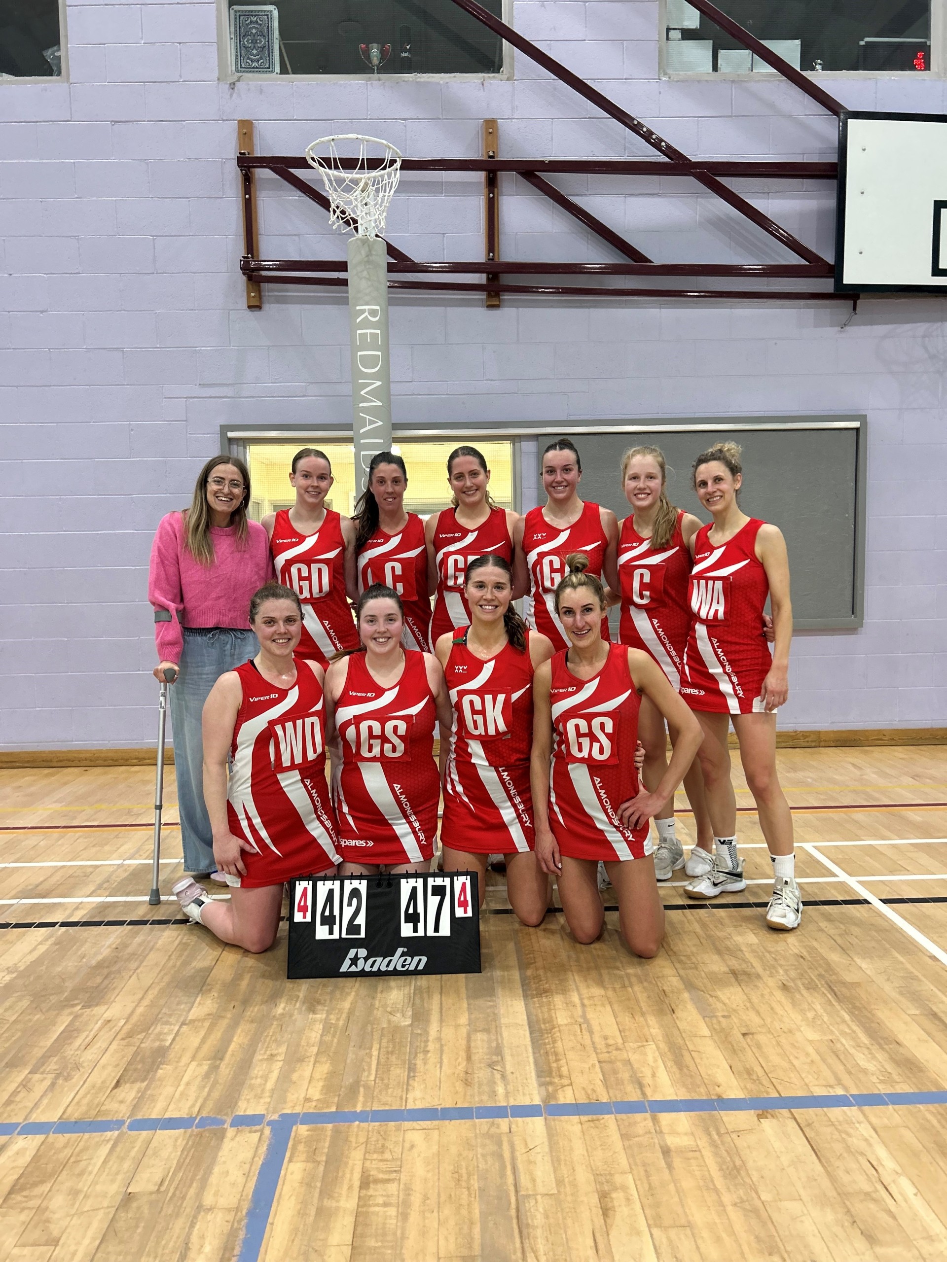 Eleven women pose in a gym, ten wearing red and white netball uniforms, and one in casual clothes with crutches. They stand behind a scoreboard reading 42 and 47 below a netball hoop.