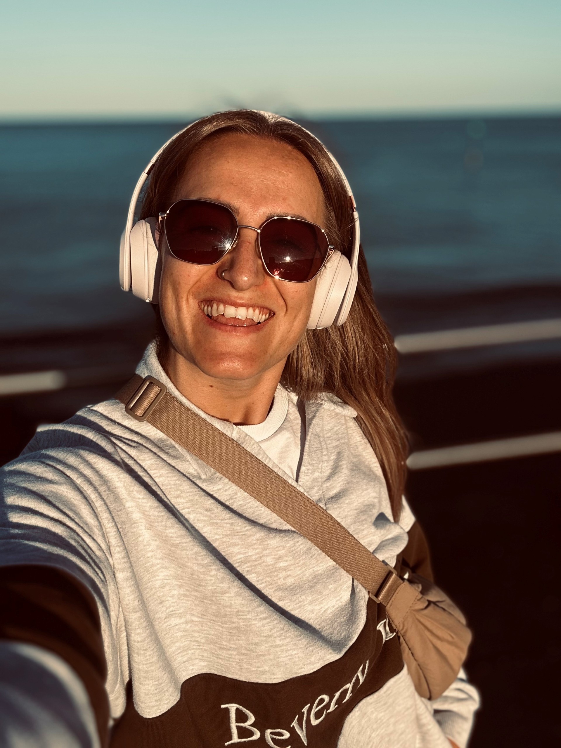 A woman wearing sunglasses and white headphones smiles while taking a selfie outdoors near a body of water, with sunlight illuminating her face. She is wearing a light gray sweatshirt and a beige crossbody bag.