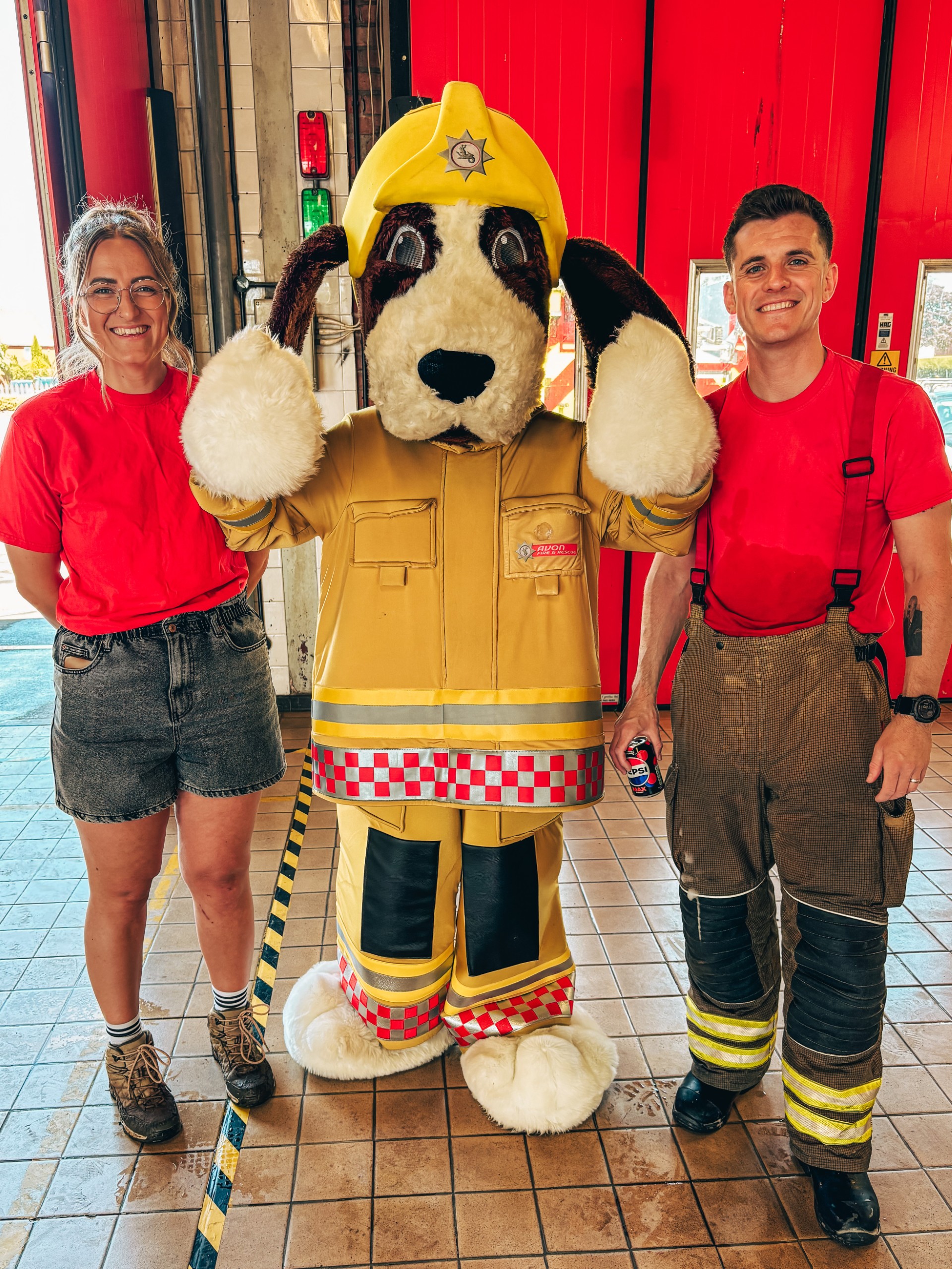 Two people in red shirts stand beside a large dog mascot dressed as a firefighter in a yellow uniform and helmet, posing for a photo inside a fire station with bright red doors in the background.