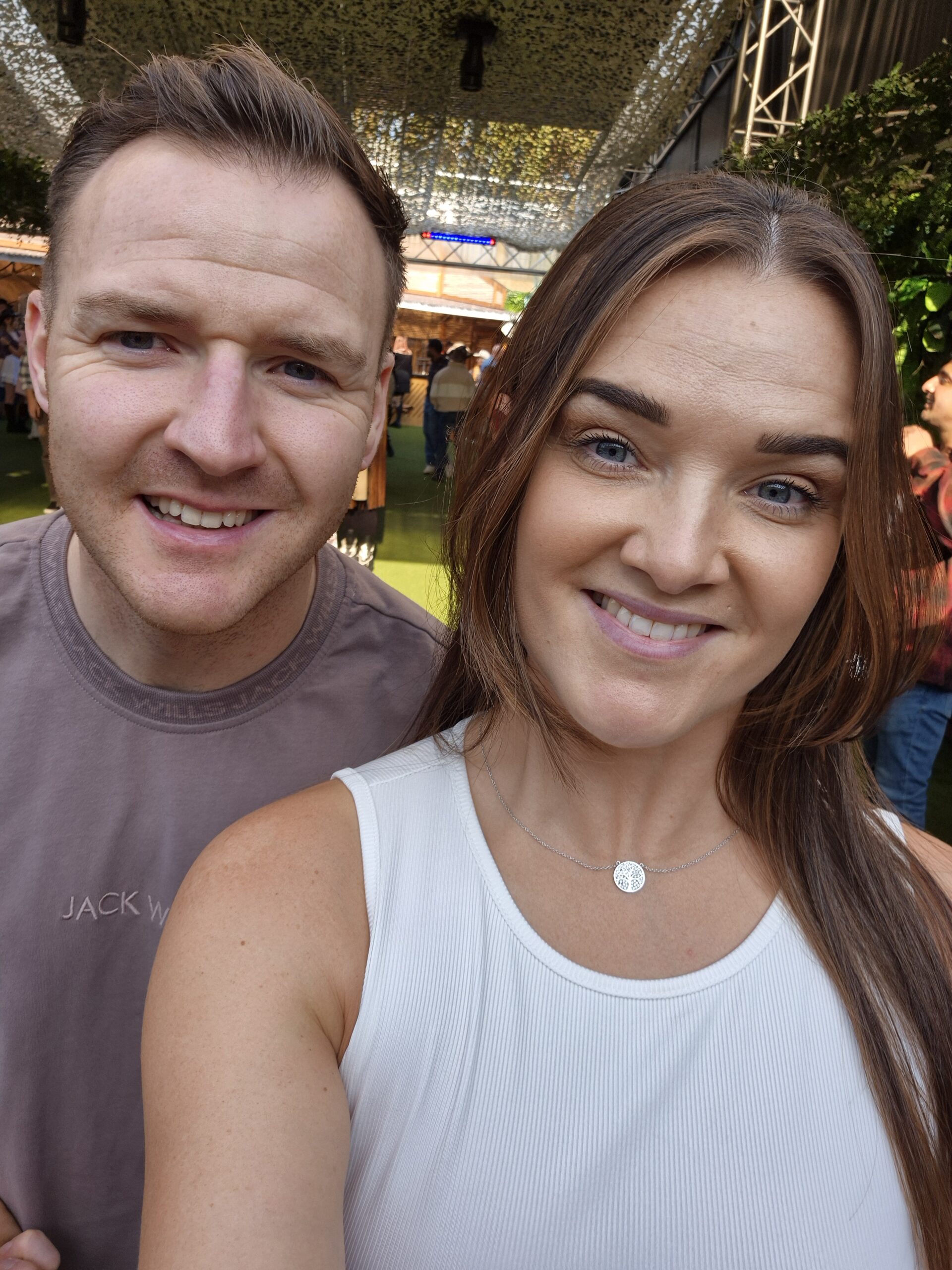 A man and woman smile for a selfie at an outdoor event with a green canopy above and people in the background. The woman wears a white top, and the man wears a mauve T-shirt.