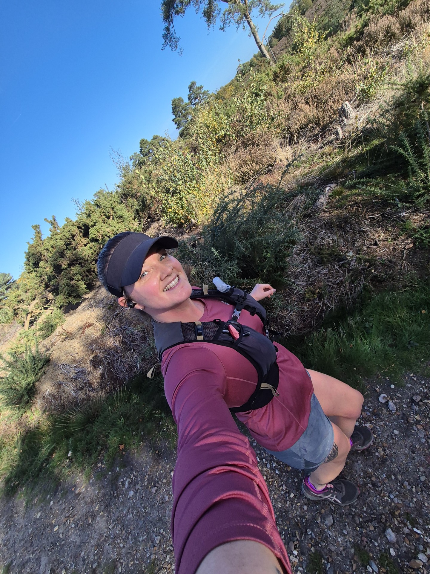 A woman wearing a cap, maroon long-sleeve shirt, and shorts takes a smiling selfie while hiking on a sunny day, with greenery and shrubs in the background.