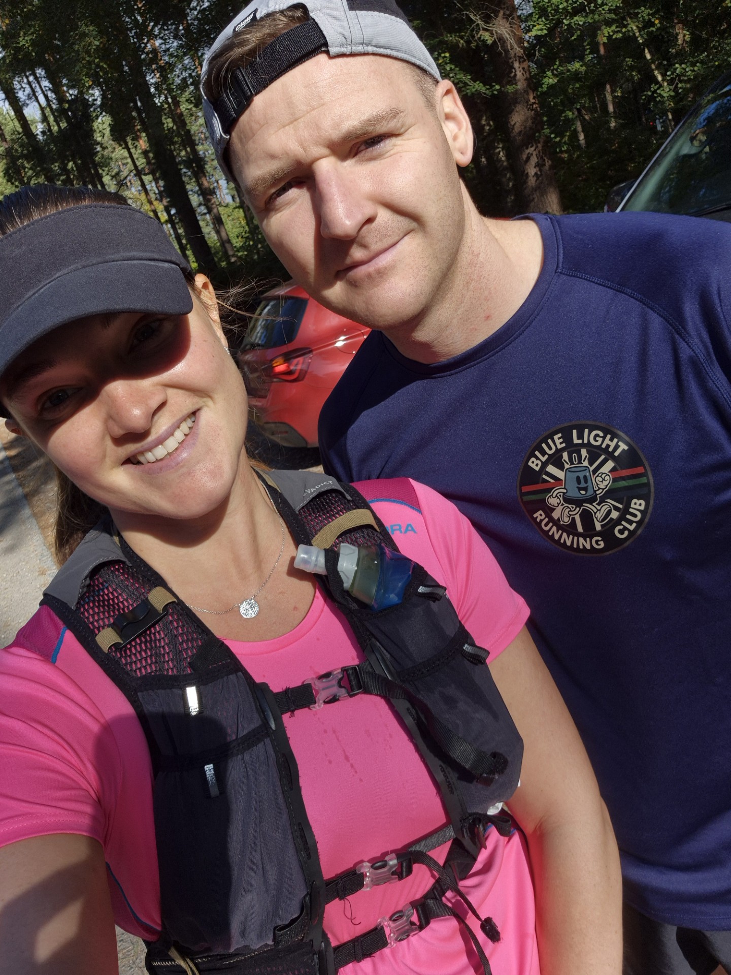 A woman in a pink running shirt and cap takes a selfie with a man in a blue "Blue Light Running Club" shirt outdoors on a sunny day, with trees and parked cars in the background.