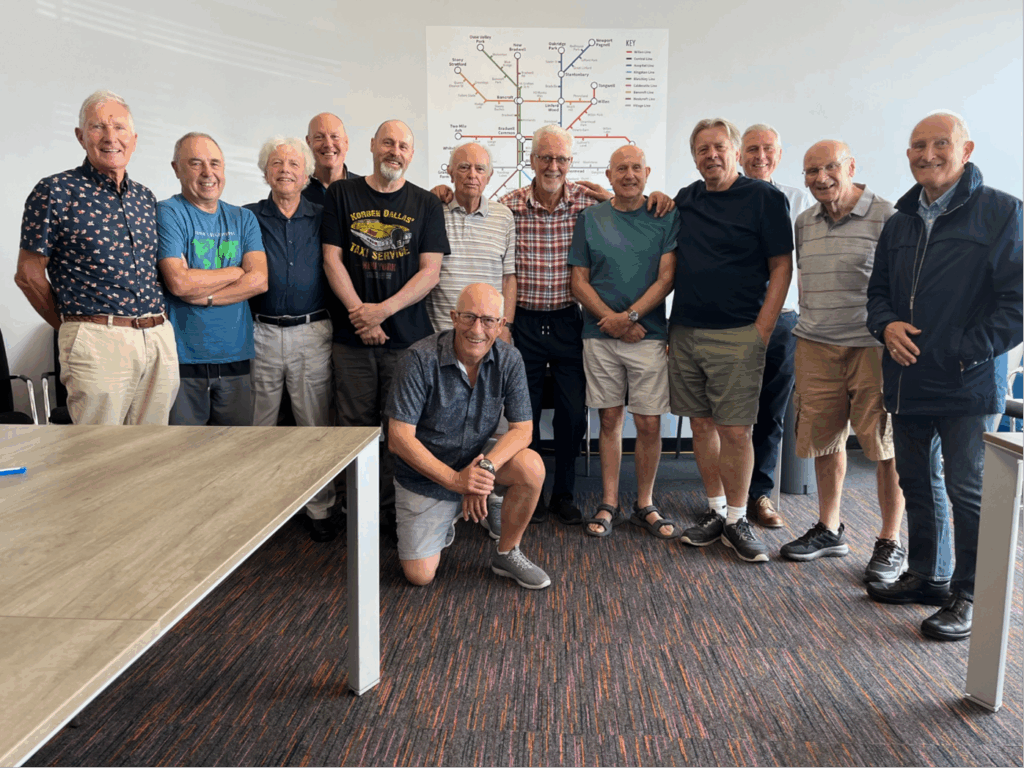A group of thirteen older men pose and smile for a photo in a room with tables and a colorful map on the wall behind them. One man kneels in front while the others stand close together.