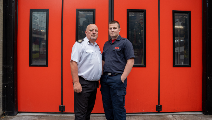 Two men wearing fire service uniforms stand side by side in front of large red doors with vertical windows, looking directly at the camera.