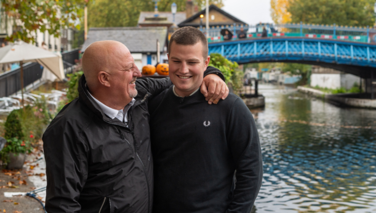 Two men stand by a canal, smiling and laughing together. One has his arm around the other. A blue bridge and trees with autumn leaves are visible in the background.