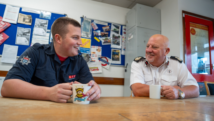 Two men in fire service uniforms sit at a table, smiling and holding mugs, in a room with noticeboards covered in posters and papers behind them.
