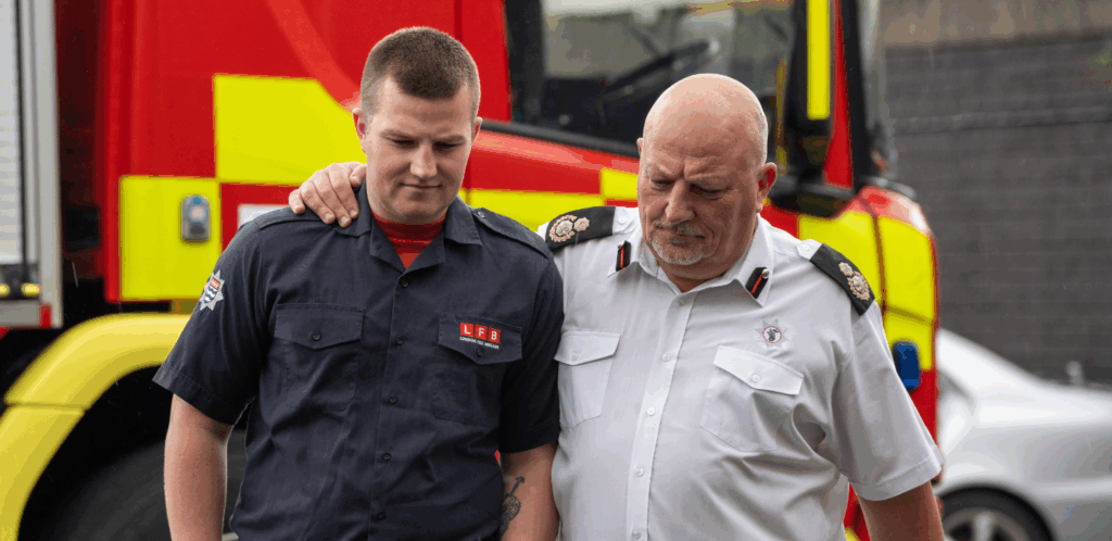 Two firefighters, one in a dark uniform and the other in a white shirt, walk together with serious expressions. The older man has his arm around the younger man’s shoulders. A fire truck is visible in the background.