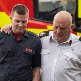 Two firefighters, one in a dark uniform and the other in a white shirt, walk together with serious expressions. The older man has his arm around the younger man’s shoulders. A fire truck is visible in the background.