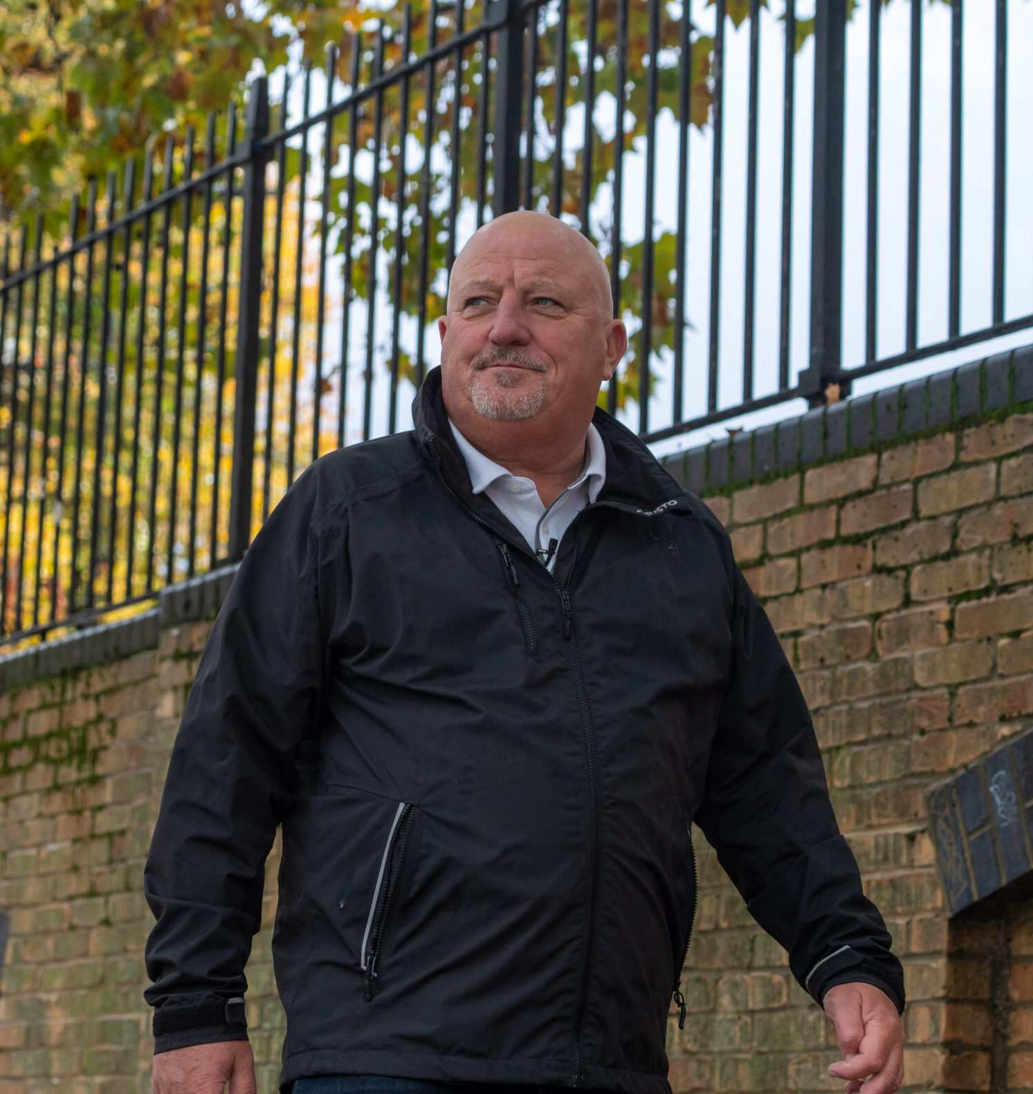 Two men walking outdoors by a brick wall and a metal fence, with trees featuring green and yellow leaves in the background. Both are dressed in dark clothing and appear to be engaged in conversation.