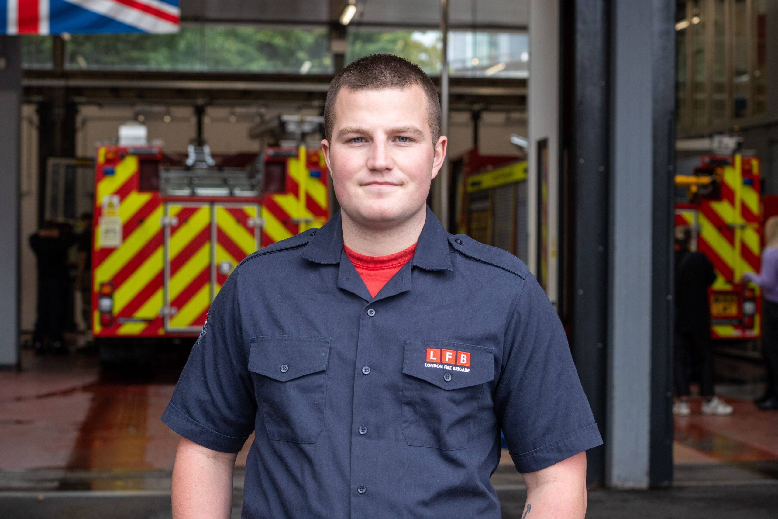 A firefighter in uniform stands outside a fire station, with fire trucks visible in the background. The person faces the camera and smiles slightly.
