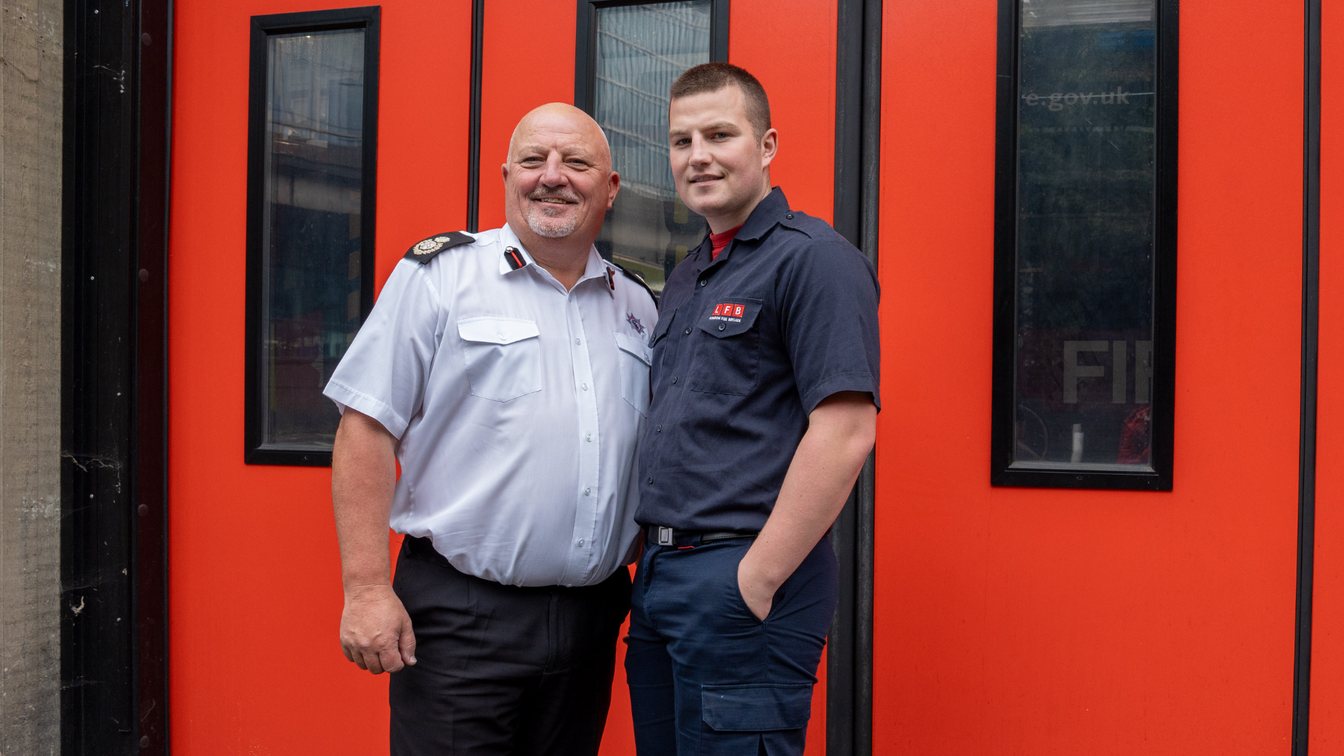 Two firefighters, one older in a white uniform shirt and one younger in a navy uniform, stand smiling together in front of bright red fire station doors.
