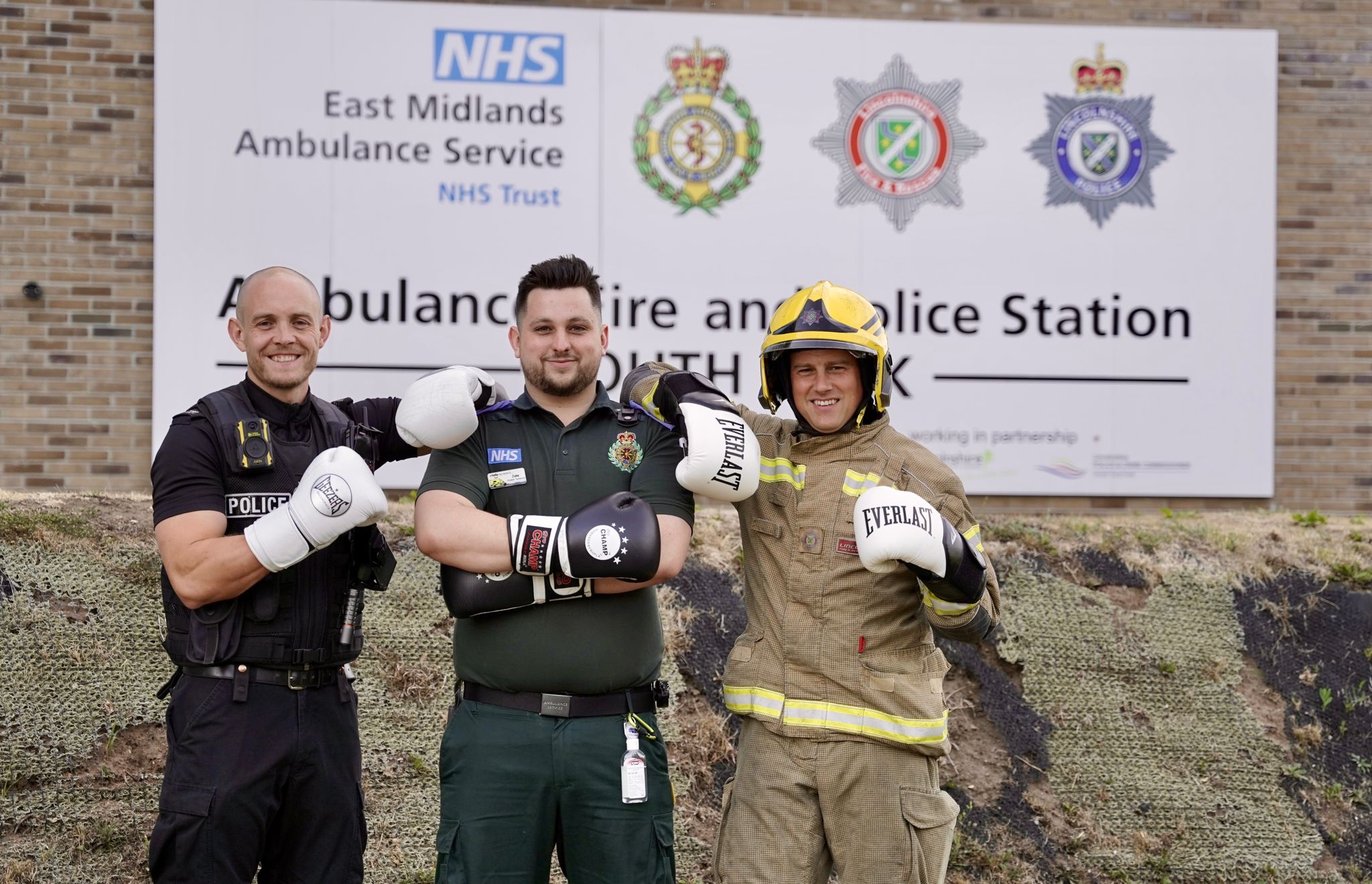 Three emergency service workers—a police officer, a paramedic, and a firefighter—stand smiling with boxing gloves on in front of an Ambulance and Police Station sign.