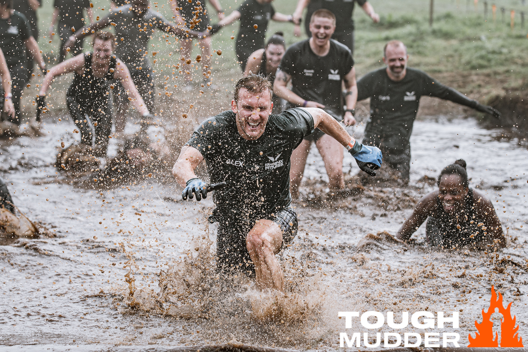 A group of people covered in mud, splashing through a muddy water pit during a Tough Mudder obstacle race. The participants appear energetic and determined, with the Tough Mudder logo at the bottom right.