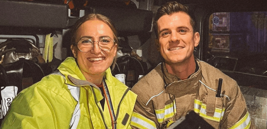 Two firefighters, one wearing a yellow jacket and glasses and the other in tan gear, smile at the camera while sitting inside a fire truck.