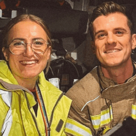 Two firefighters, one wearing a yellow jacket and glasses and the other in tan gear, smile at the camera while sitting inside a fire truck.