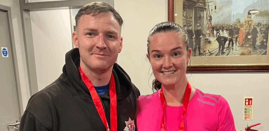 Two people wearing medals smile for the camera indoors. The man is in a black hoodie and the woman in a pink top. Behind them is a framed painting of firefighters and emergency responders in action.