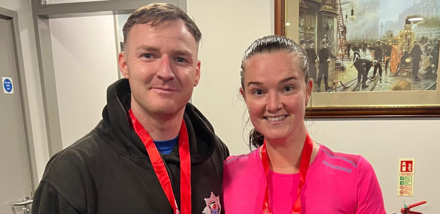 Two people wearing medals smile for the camera indoors. The man is in a black hoodie and the woman in a pink top. Behind them is a framed painting of firefighters and emergency responders in action.