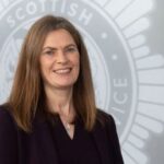 A woman with straight, shoulder-length brown hair, wearing a dark blazer, stands smiling in front of a wall with the Scottish Fire and Rescue Service emblem in the background.