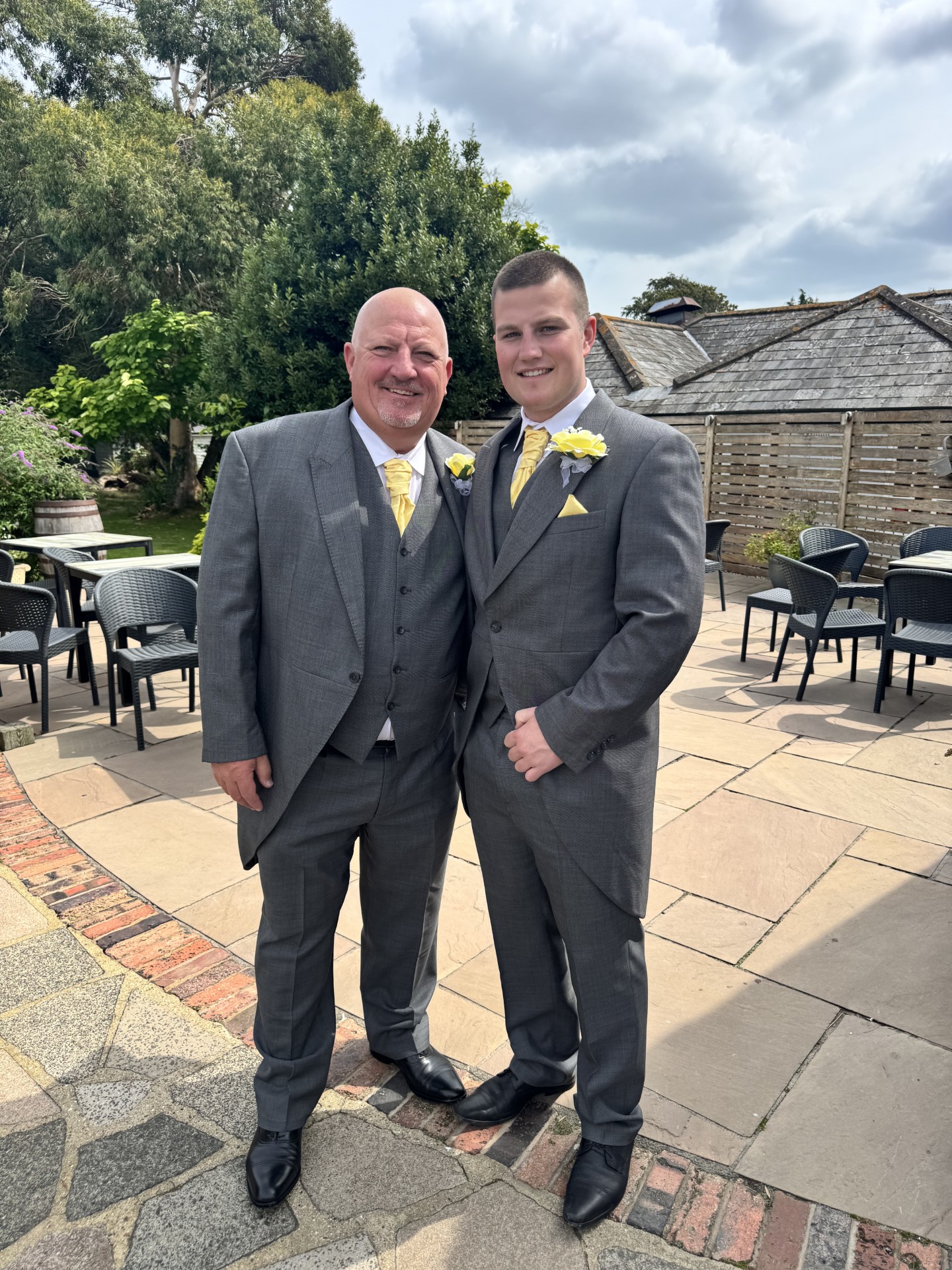 Two men dressed in matching grey suits with yellow ties and white rose boutonnieres smile and pose together outside on a patio with chairs, tables, and greenery in the background.