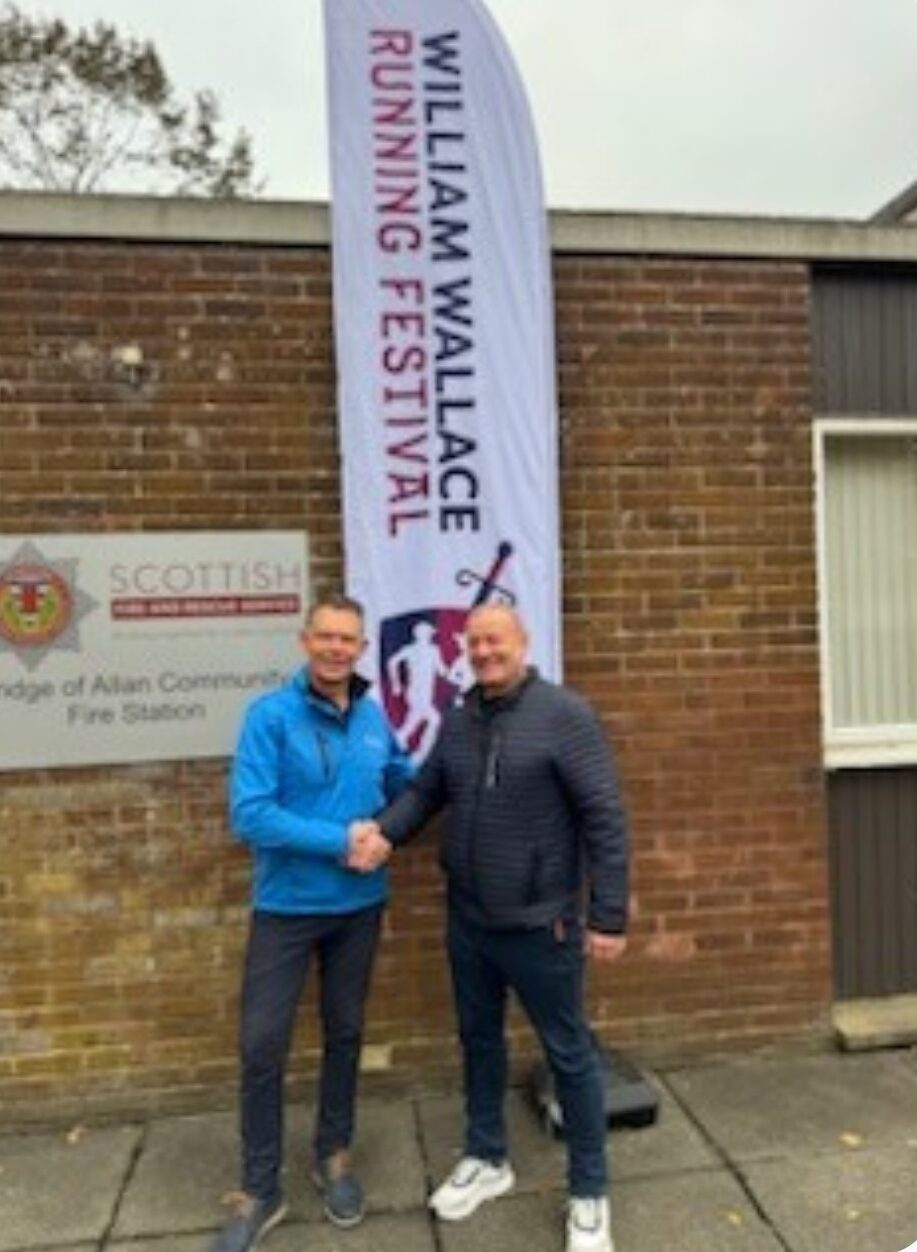 Two men are shaking hands and smiling in front of a "William Wallace Running Festival" banner, standing outside a building with a "Scottish" sign on the wall.
