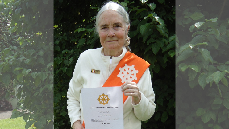 An older woman with gray hair stands outdoors in front of green foliage, wearing a white top and an orange sash with a white wheel symbol. She holds a certificate and looks at the camera.