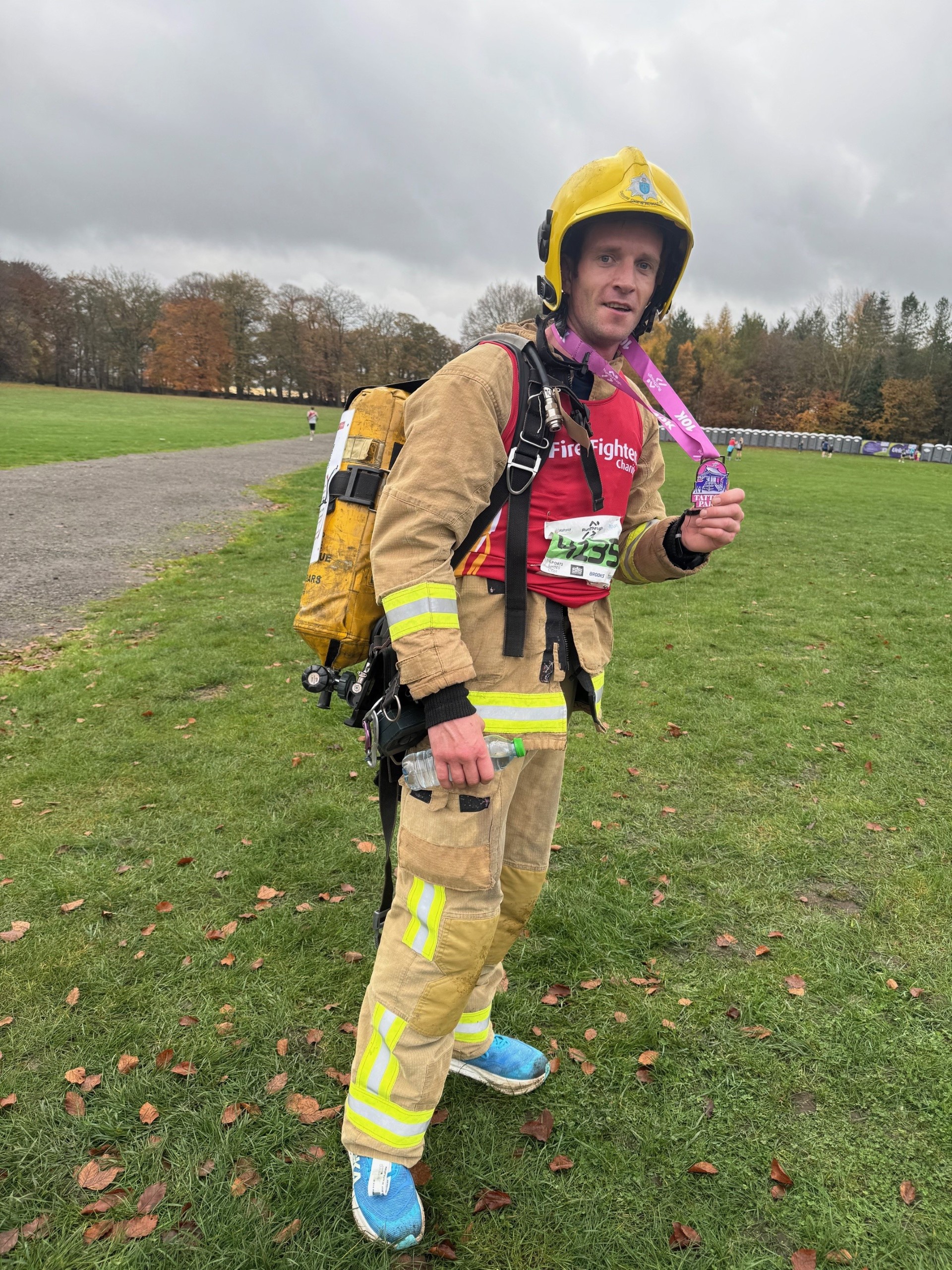A man in full firefighter gear stands on a grassy field, holding a medal around his neck. He is wearing a yellow helmet, reflective suit, backpack, and blue running shoes. Trees and a cloudy sky are in the background.