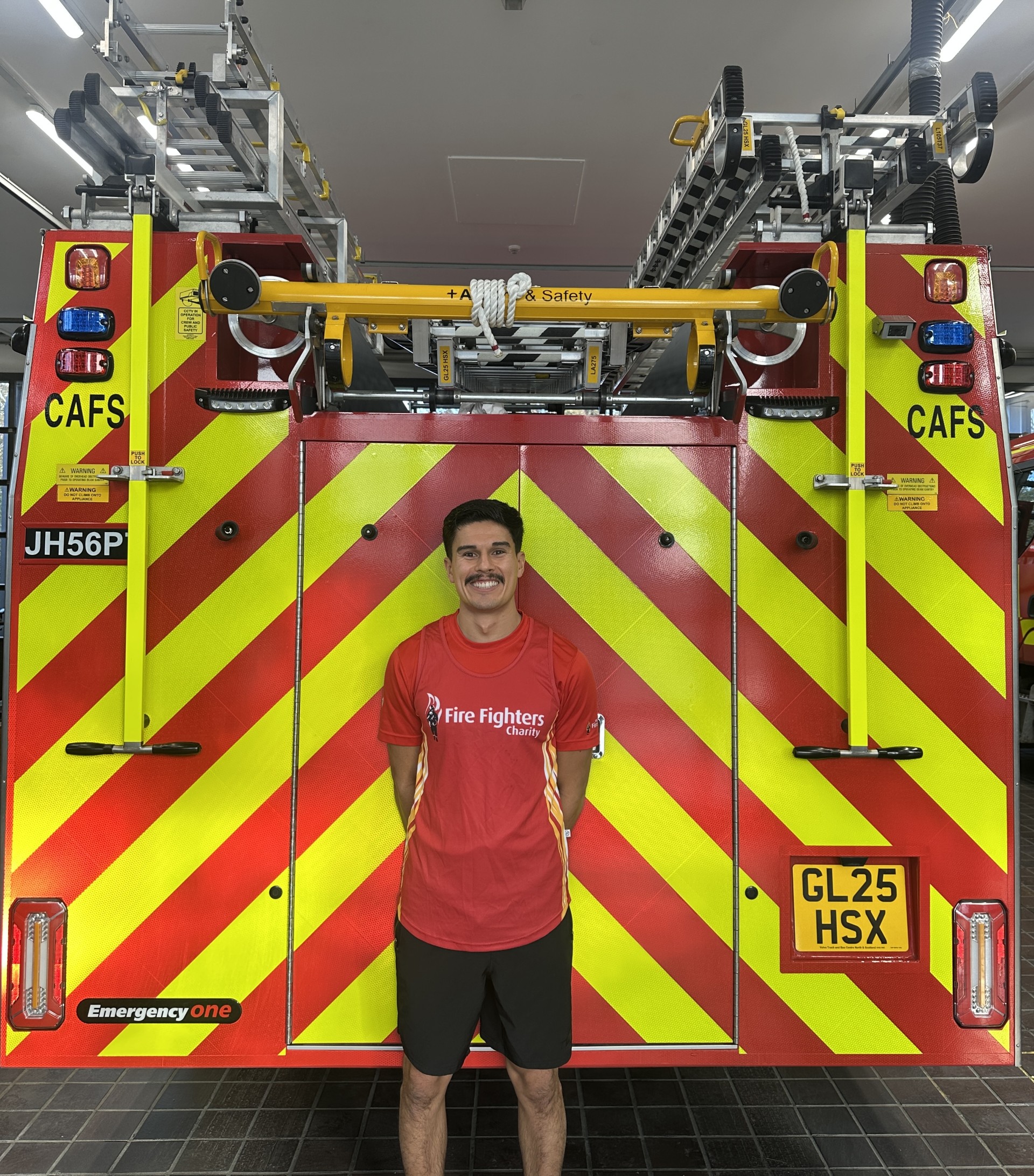 A man in a red "Fire Fighters" t-shirt stands smiling in front of a fire truck with yellow and red diagonal stripes, ladders on top, and license plates reading "JH56P" and "GL25 HSX.