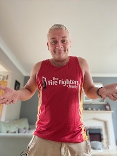 A man with short blonde hair, wearing a red "The Fire Fighters Charity" tank top and beige shorts, stands indoors with his arms raised and shrugged, smiling slightly. A fireplace and sofa are visible in the background.