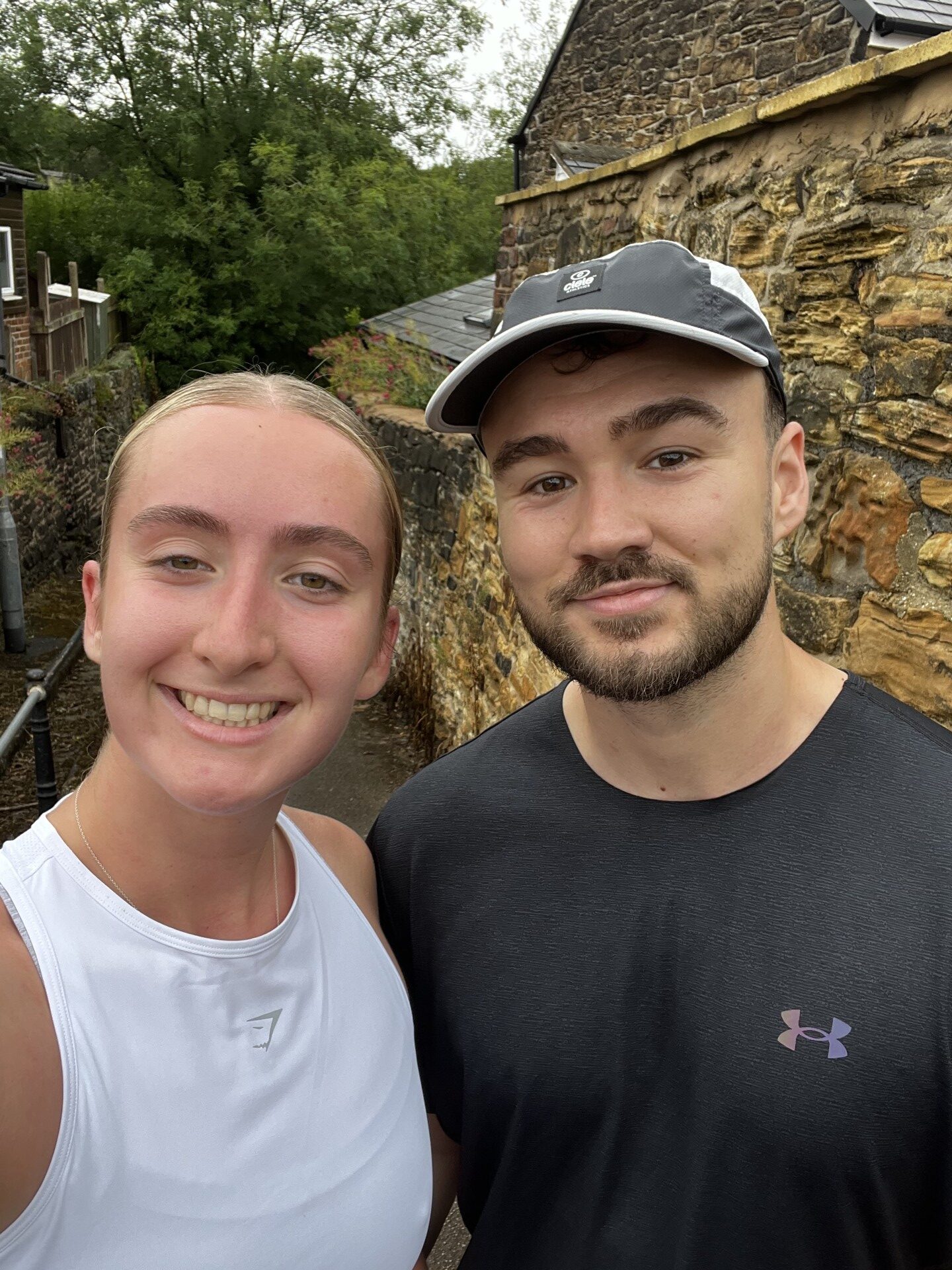 Two people smiling at the camera outdoors, standing close together. One is wearing a grey cap and black shirt, the other is in a white tank top. A stone wall and greenery are visible in the background.