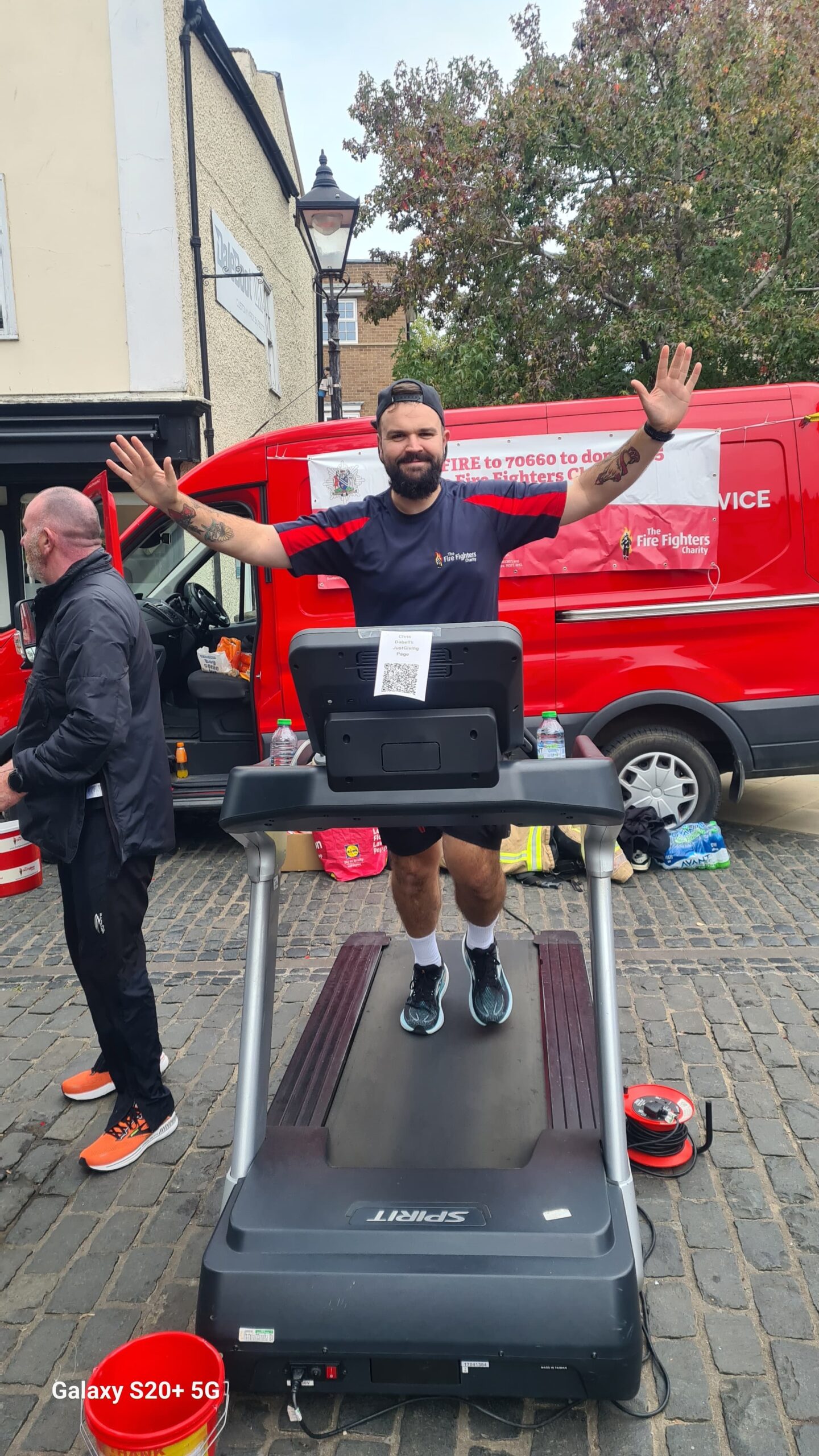 A man with a beard stands on a treadmill outdoors, smiling with arms raised. Behind him, a red van and another man are visible. The setting is a cobblestone street with buildings and trees in the background.