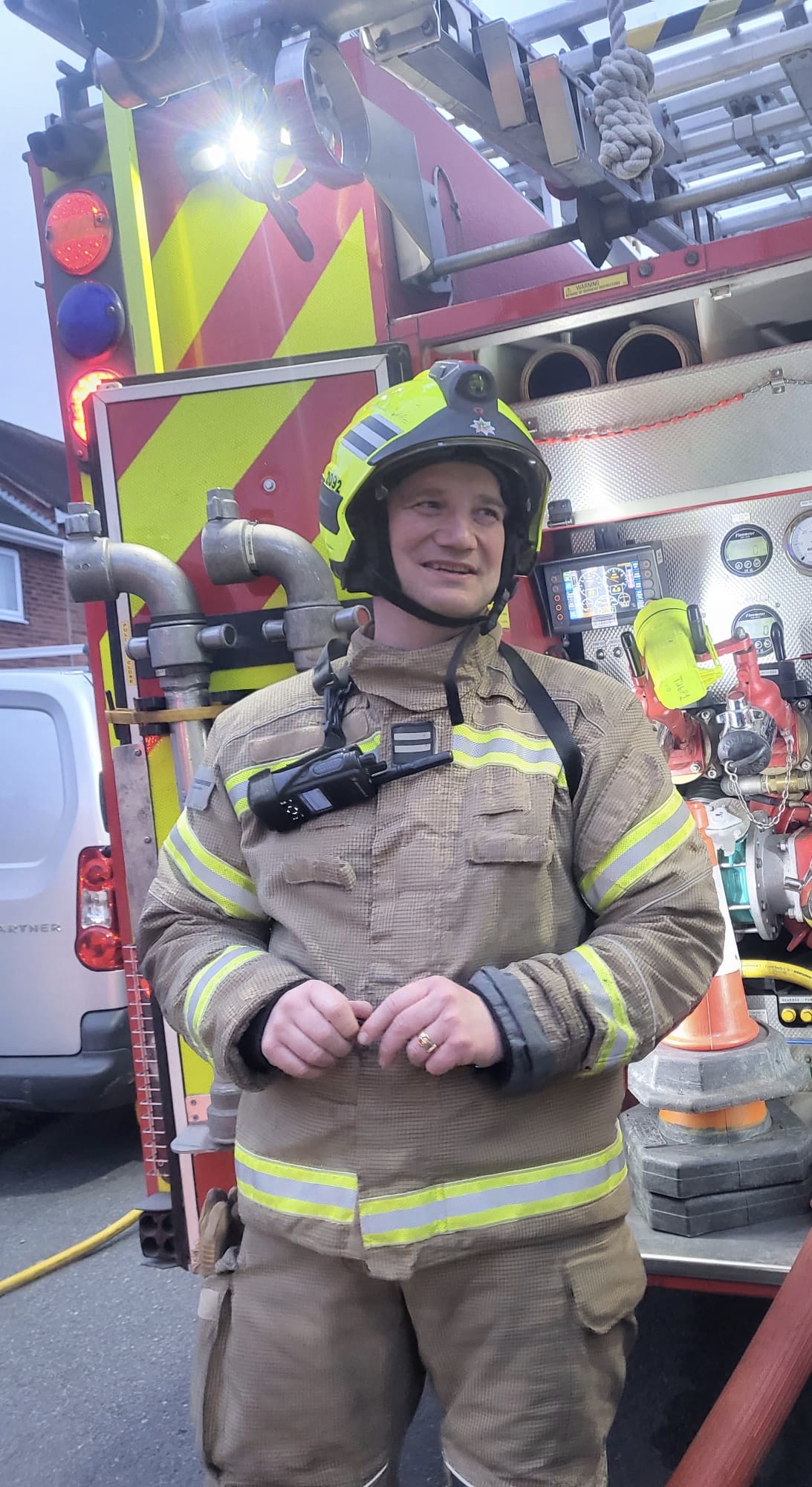 A firefighter in full protective gear and helmet stands smiling in front of a fire engine, with equipment and hoses visible in the background.